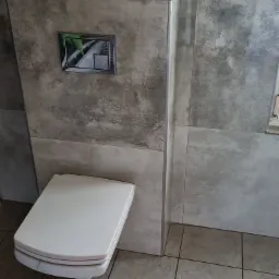 Kompakt WC z ukrytą spłuczką, obudowany płytami imitującymi beton, z chromowanym przyciskiem spłukującym.