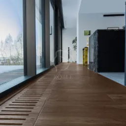 Wnętrze nowoczesnego domu z brązową podłogą z paneli, dużym oknem panoramicznym z widokiem na zimowy krajobraz i grzejnikiem kanałowym w podłodze.