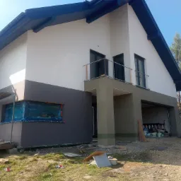 Dom jednorodzinny w trakcie ocieplania elewacji: dolna część w kolorze grafitowym, górna biała, okno zabezpieczone folią, widoczne rusztowania i materiały budowlane.