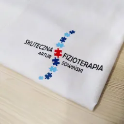 Biała koszulka z nadrukiem logo 'Skuteczna Fizjoterapia Artur Sowiński', stylizowanego na krzyż z puzzli w kolorach niebieskim i czerwonym, leżąca na drewnianym blacie.