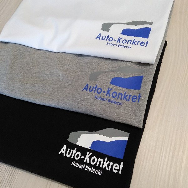 Stos trzech złożonych koszulek: białej, szarej i czarnej, każda z nadrukowanym logo firmy 'Auto-Konkret Hubert Bielecki' w kolorach szarym, białym i niebieskim.