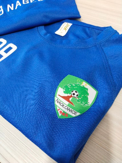 Niebieska koszulka sportowa z naszywką klubu sportowego Nagłowice, widoczny fragment napisu na drugiej koszulce w tle.