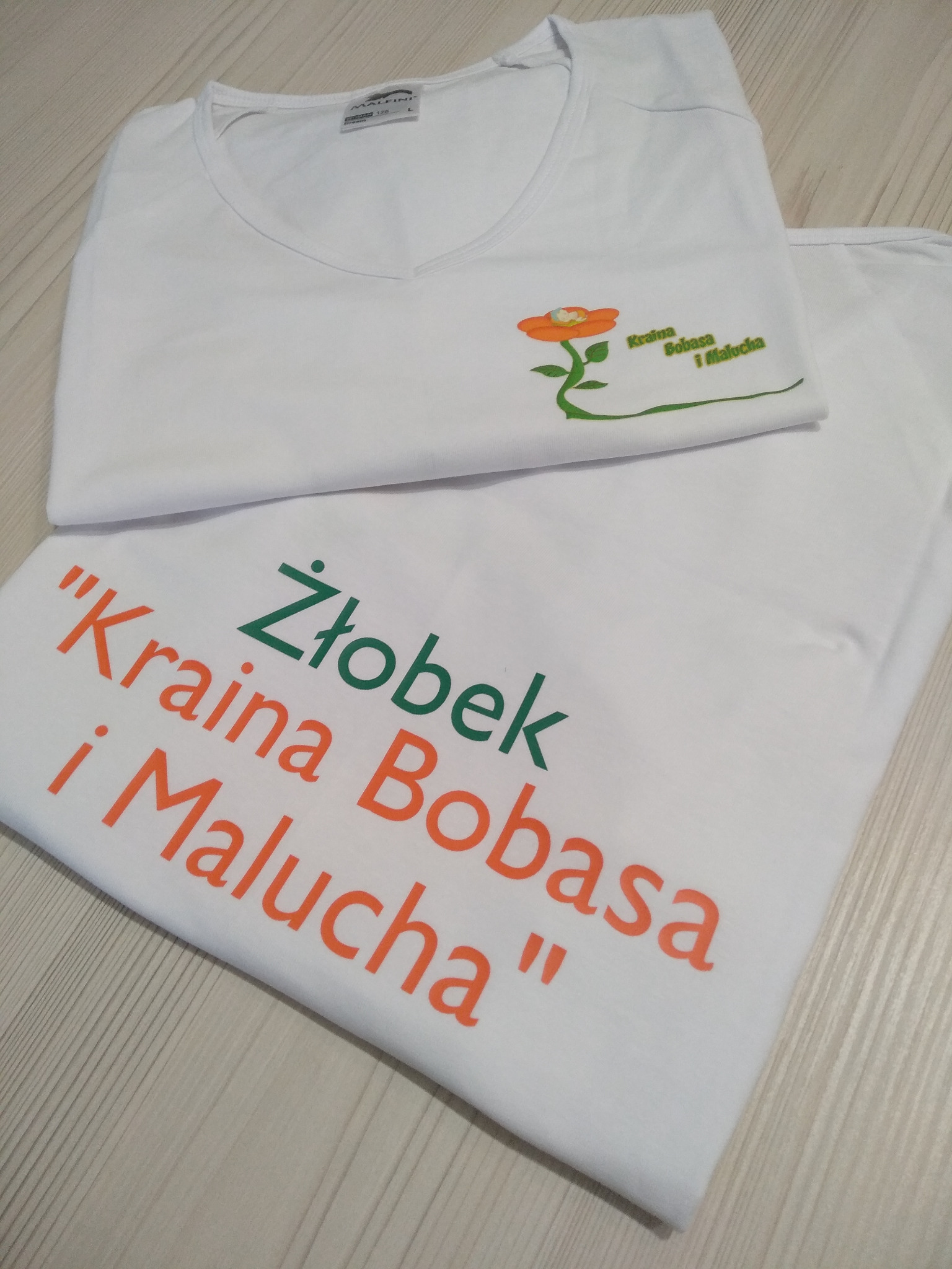 Dwa białe T-shirty z nadrukami: jeden z pomarańczowym kwiatem i zielonym napisem 'Kraina Bobasa i Malucha', drugi z zielonym napisem 'Żłobek' i pomarańczowym napisem 'Kraina Bobasa i Malucha'.