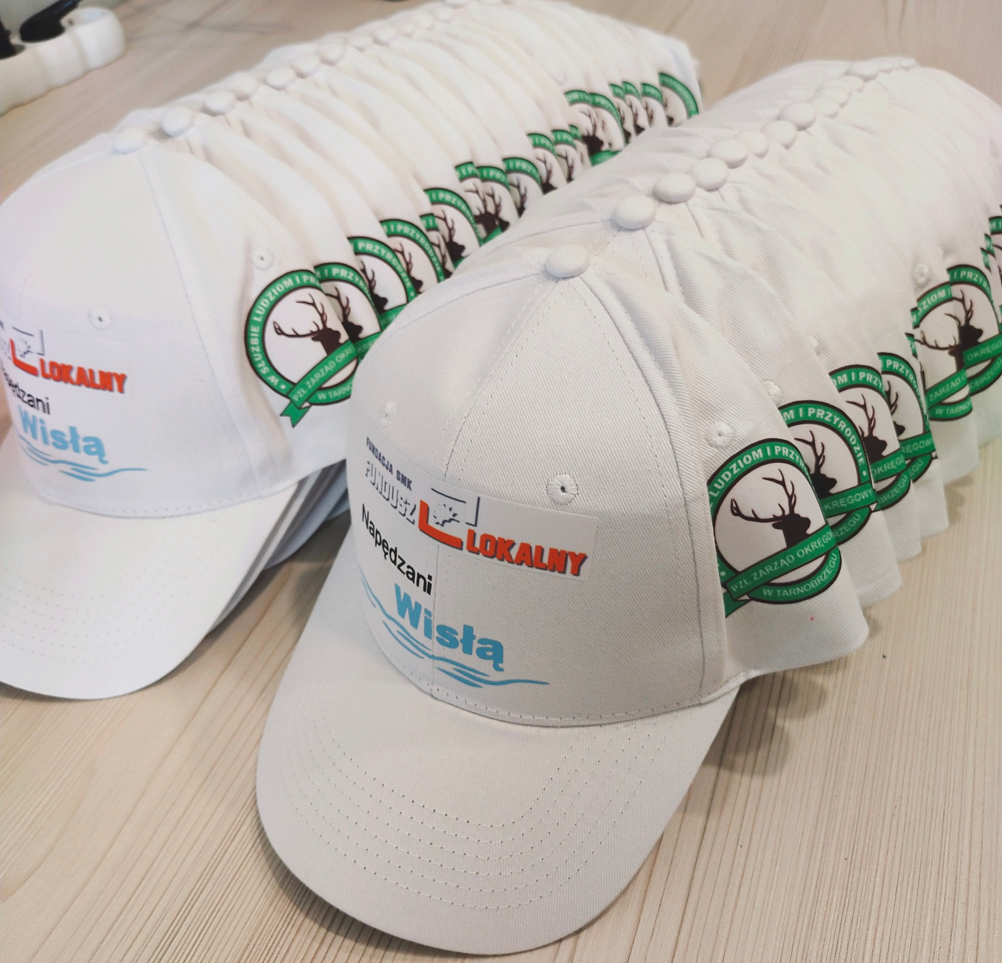 Białe czapki z nadrukiem logo Fundacji SMK Fundusz Lokalny Napędzani Wisłą i logotypem PZŁ Zarząd Okręgowy w Tarnobrzegu, ułożone w rzędzie na drewnianym podłożu.