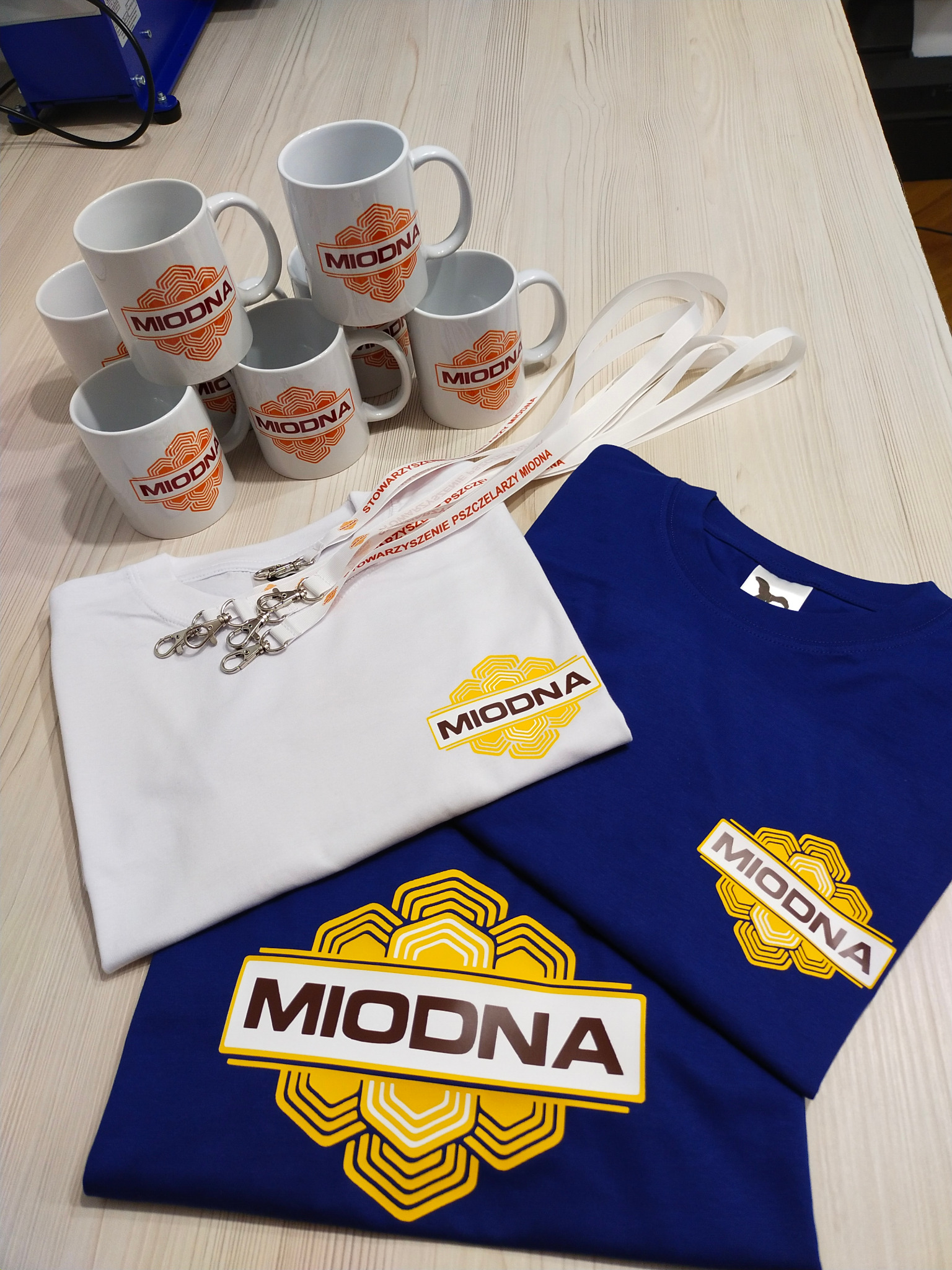 Kolekcja gadżetów reklamowych z nadrukowanym logo 'Miodna' na białych kubkach, białej i granatowej koszulce oraz smyczach z karabińczykami, ułożonych na jasnym drewnianym blacie.