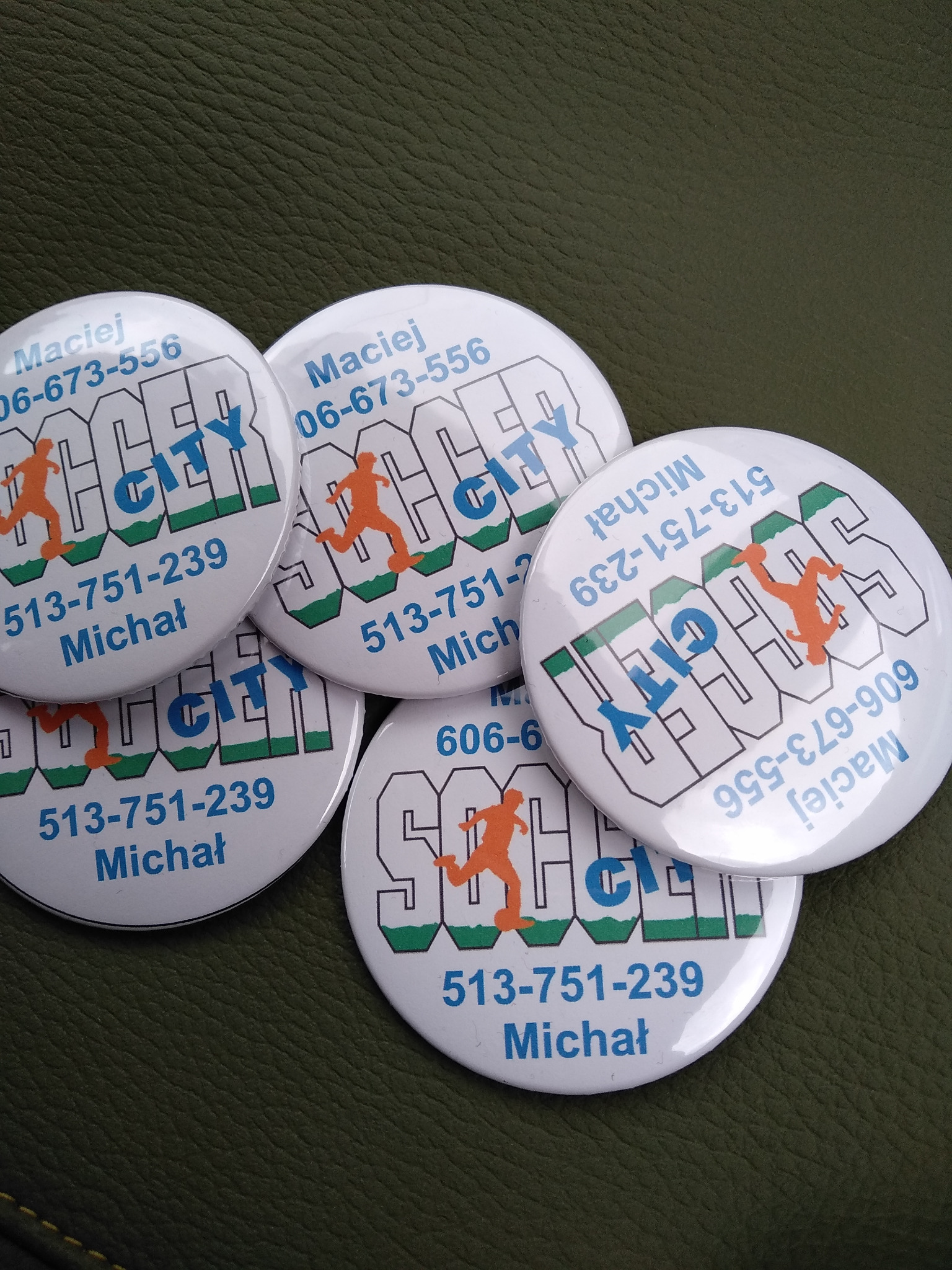 Stos okrągłych przypinek z nadrukowanym logo 'Soccer City', numerami telefonów i imionami Maciej i Michał, leżących na teksturowanym, ciemnozielonym tle.