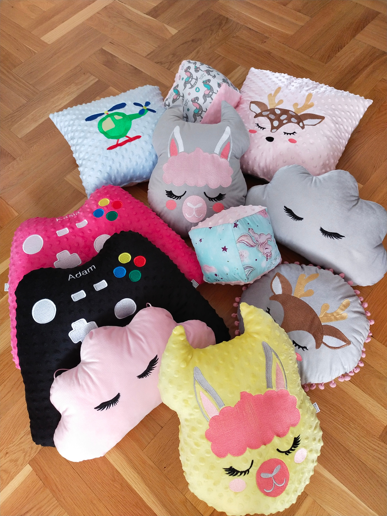 Kolekcja dekoracyjnych poduszek w różnych kształtach i kolorach, w tym poduszki w kształcie kontrolera do gier z imionami Adam i Kasia, lama, chmury, jelenia i helikoptera, leżące na drewnianej...