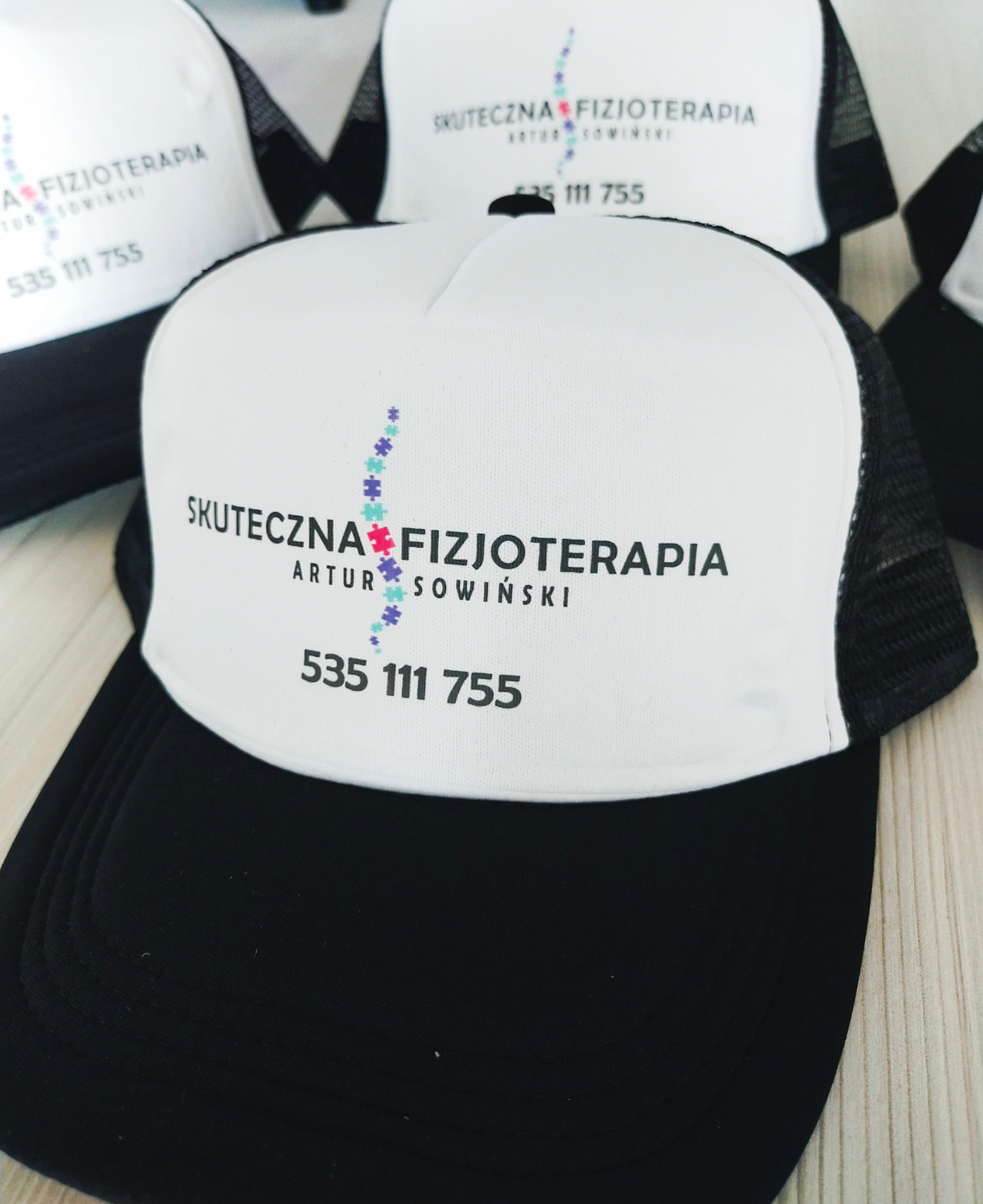 Czapki typu trucker z białym panelem frontowym i czarnym daszkiem, z nadrukowanym logo i nazwą firmy 'Skuteczna Fizjoterapia Artur Sowiński' oraz numerem telefonu.