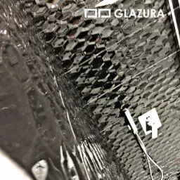 Ściana wyłożona czarnymi, heksagonalnymi płytkami z połyskiem, z zamontowaną białą baterią prysznicową i widocznym logo 'GLAZURA' w rogu.