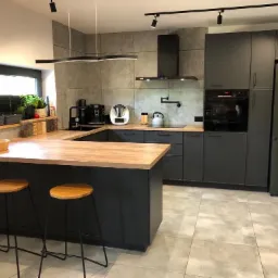 Nowoczesna kuchnia w odcieniach szarości z drewnianym blatem, oświetleniem punktowym i dwoma hokerami przy wyspie.