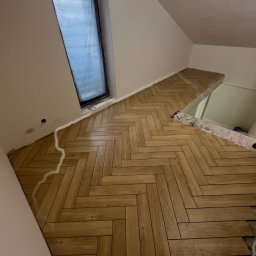 D.D. GLAZURA DAWID DZIERŻĘGA - Ukończona podłoga z drewnopodobnych paneli ułożonych w jodełkę w pokoju na poddaszu z oknem dachowym. Częściowo widoczne niedokończone fragmenty podłogi.
