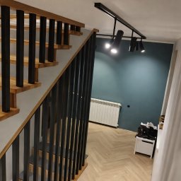D.D. GLAZURA DAWID DZIERŻĘGA - Nowoczesne schody z drewnianymi stopniami i czarną balustradą, oświetlone reflektorami. W tle ściana w odcieniach zieleni i parkiet ułożony w jodełkę.