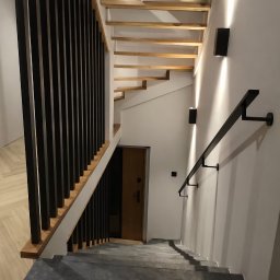 D.D. GLAZURA DAWID DZIERŻĘGA - Nowoczesne schody z drewnianymi stopniami i czarną balustradą, oświetlone kinkietami. Widok z góry na dół, z widocznymi elementami wykończenia wnętrza.