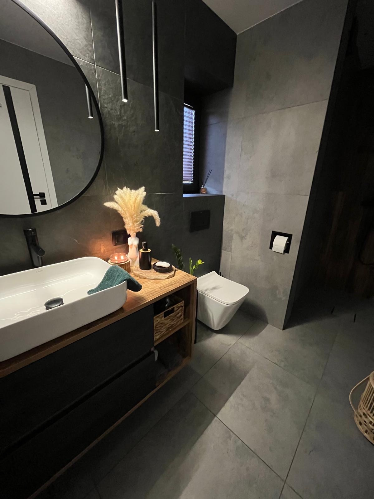 Nowoczesna łazienka w odcieniach szarości z umywalką nablatową na drewnianej szafce, akcenty dekoracyjne w postaci suszonych traw i świec, minimalistyczne oświetlenie punktowe.