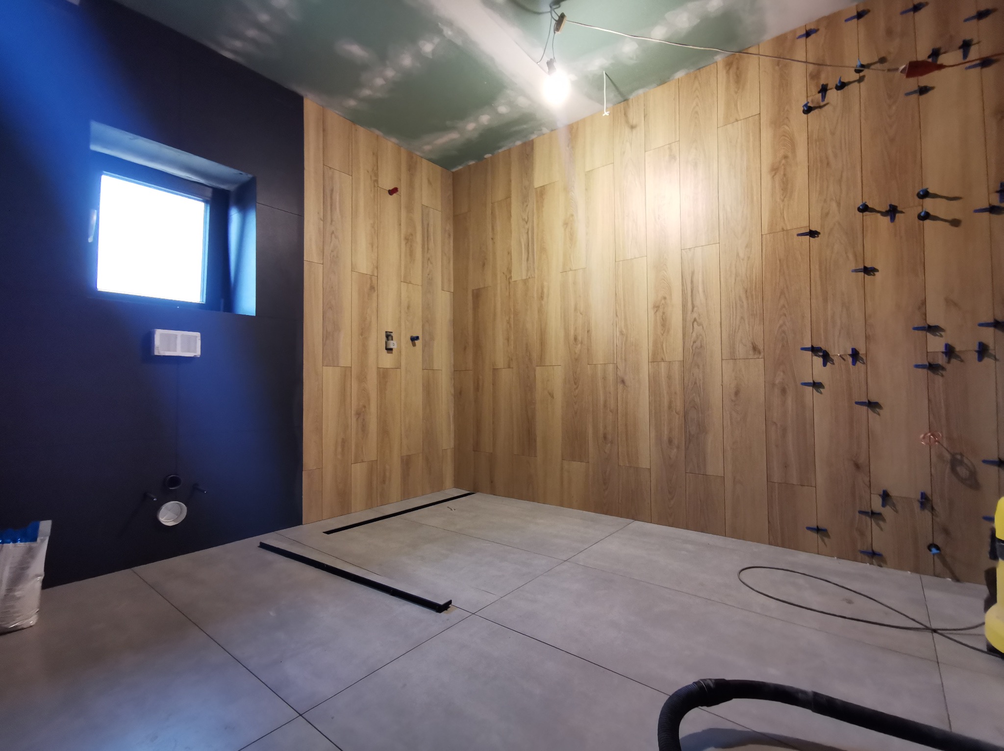 Łazienka w trakcie prac wykończeniowych: na ścianach płytki imitujące drewno z niebieskimi krzyżykami dystansowymi, na podłodze szare płytki, widoczne fragmenty instalacji i narzędzi.