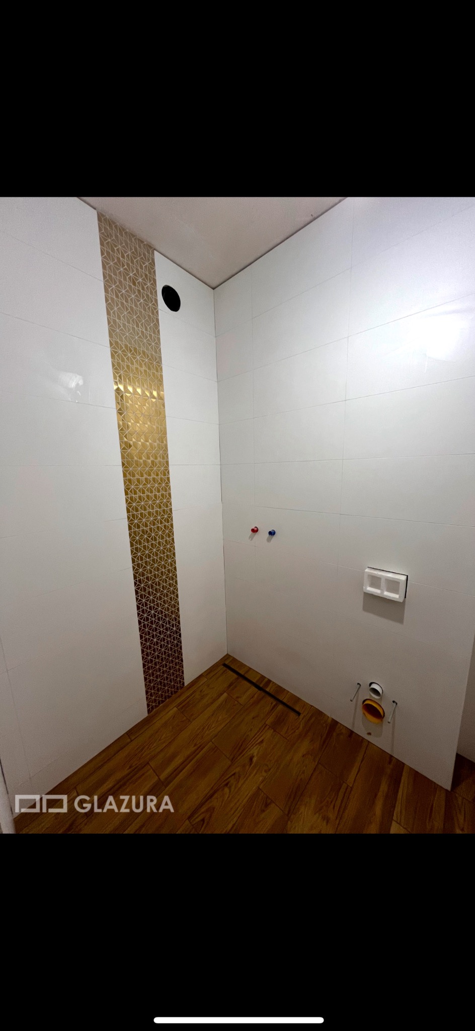 Wykończona łazienka z białymi płytkami i dekoracyjnym złotym pasem mozaiki oraz podłogą imitującą drewno, widoczne przyłącza wodne i odpływ liniowy.