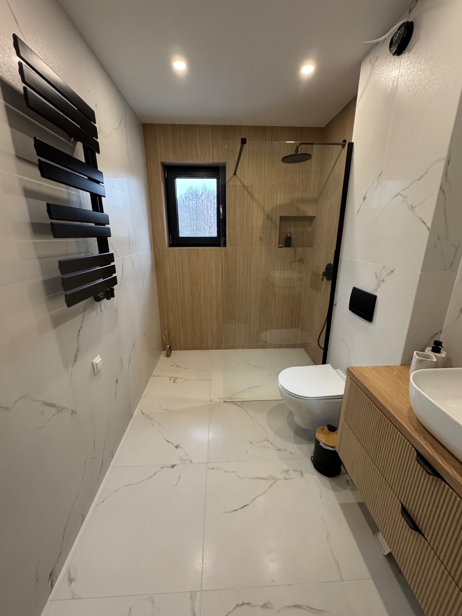 Nowoczesna łazienka z kabiną prysznicową typu walk-in, grzejnikiem drabinkowym i umywalką nablatową na drewnianej szafce. Płytki imitujące marmur i drewno.