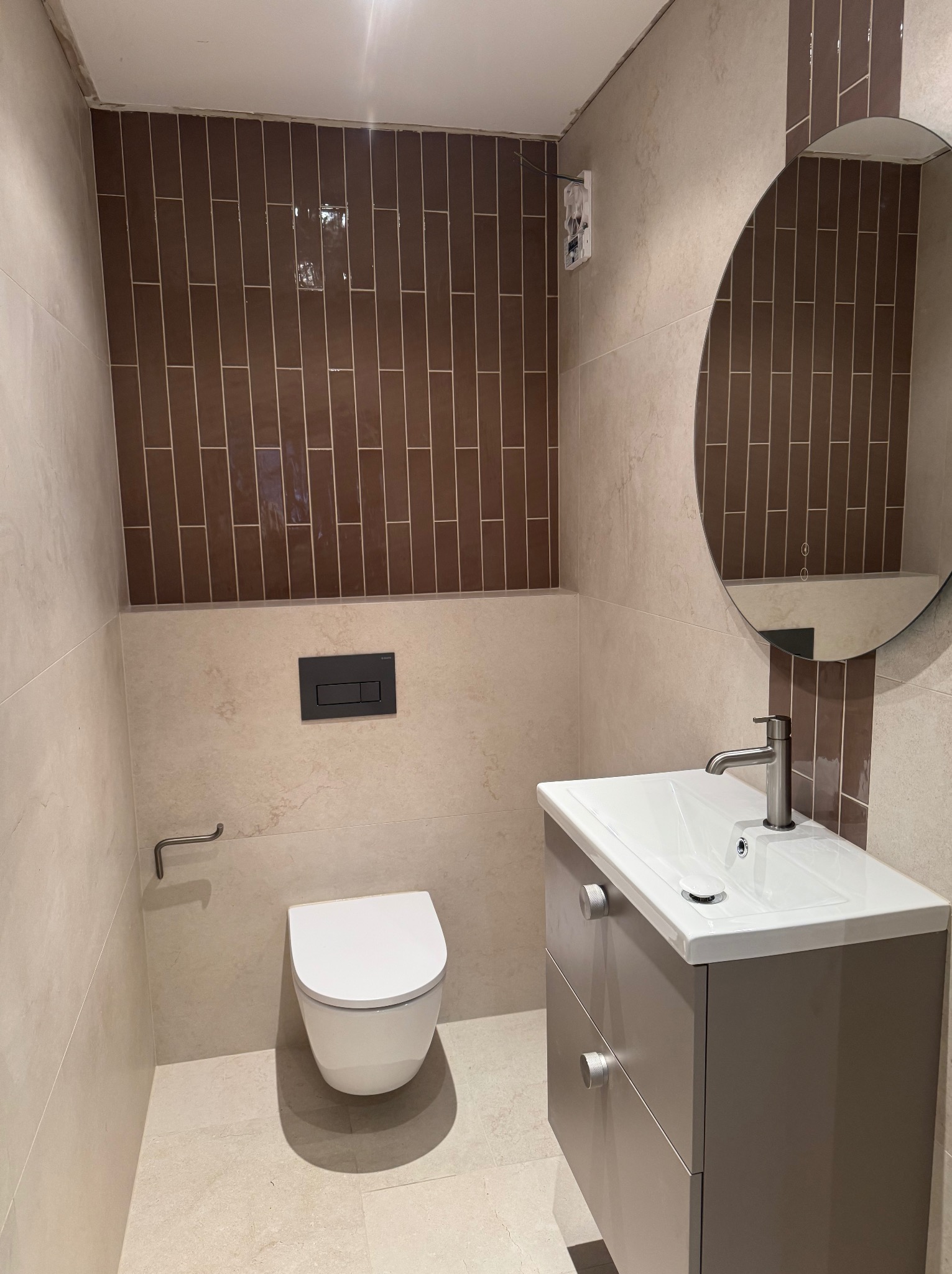 Nowoczesna łazienka po remoncie: beżowe płytki, brązowe akcenty, okrągłe lustro z podświetleniem, podwieszana toaleta i szafka z umywalką. Minimalistyczny design.