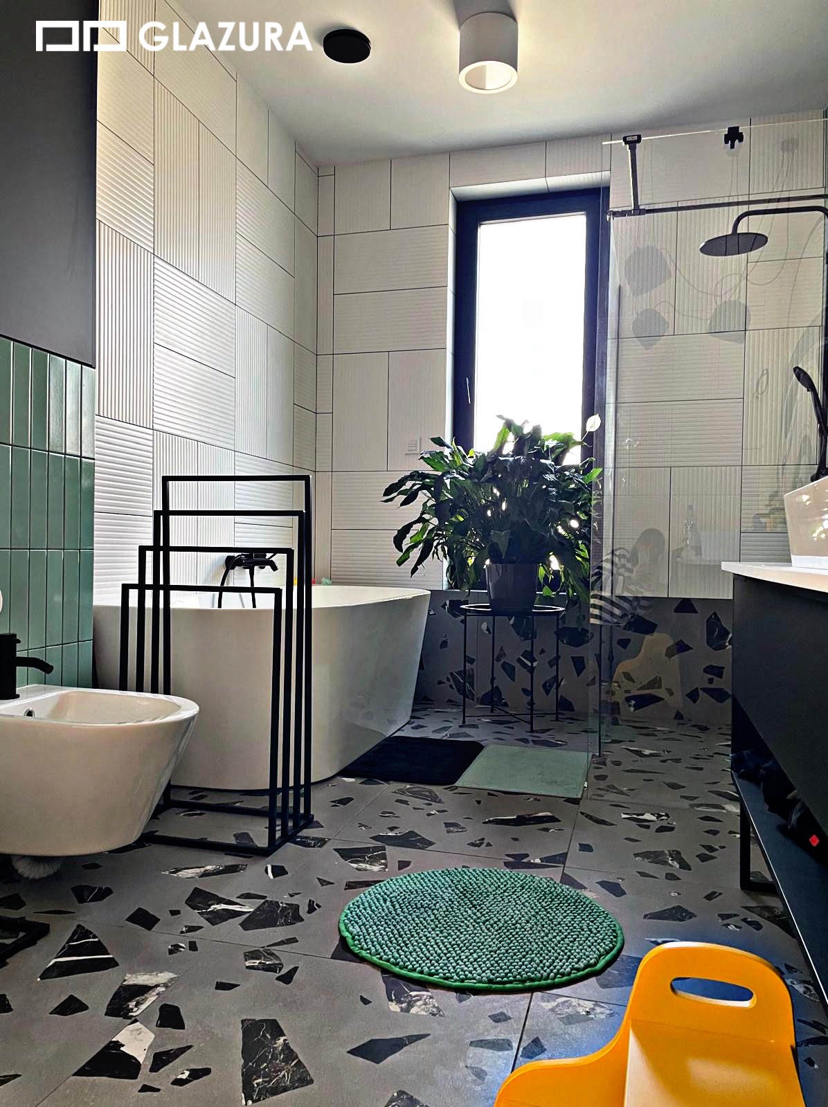 Nowoczesna łazienka z wanną wolnostojącą, bidetem i oryginalną podłogą w czarno-białe plamy. Ściany wyłożone białymi płytkami z delikatną fakturą i zielonymi akcentami.