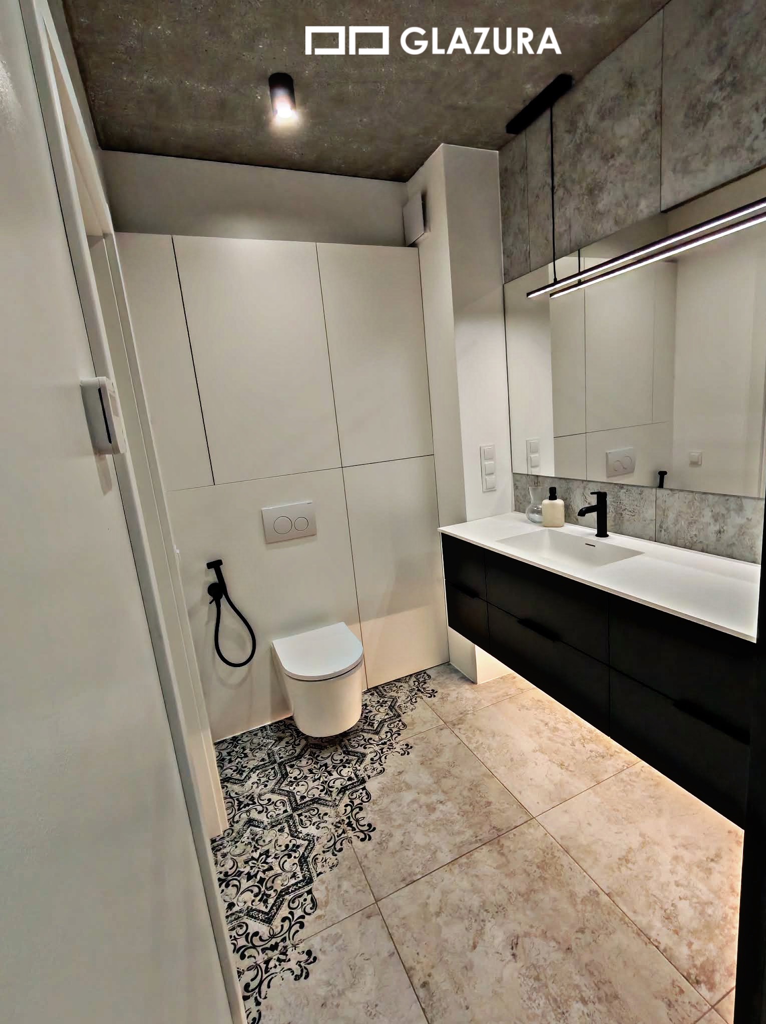 Nowoczesna łazienka z białą zabudową, czarną szafką pod umywalką i dekoracyjną podłogą z płytek w stylu vintage. Minimalistyczne oświetlenie.