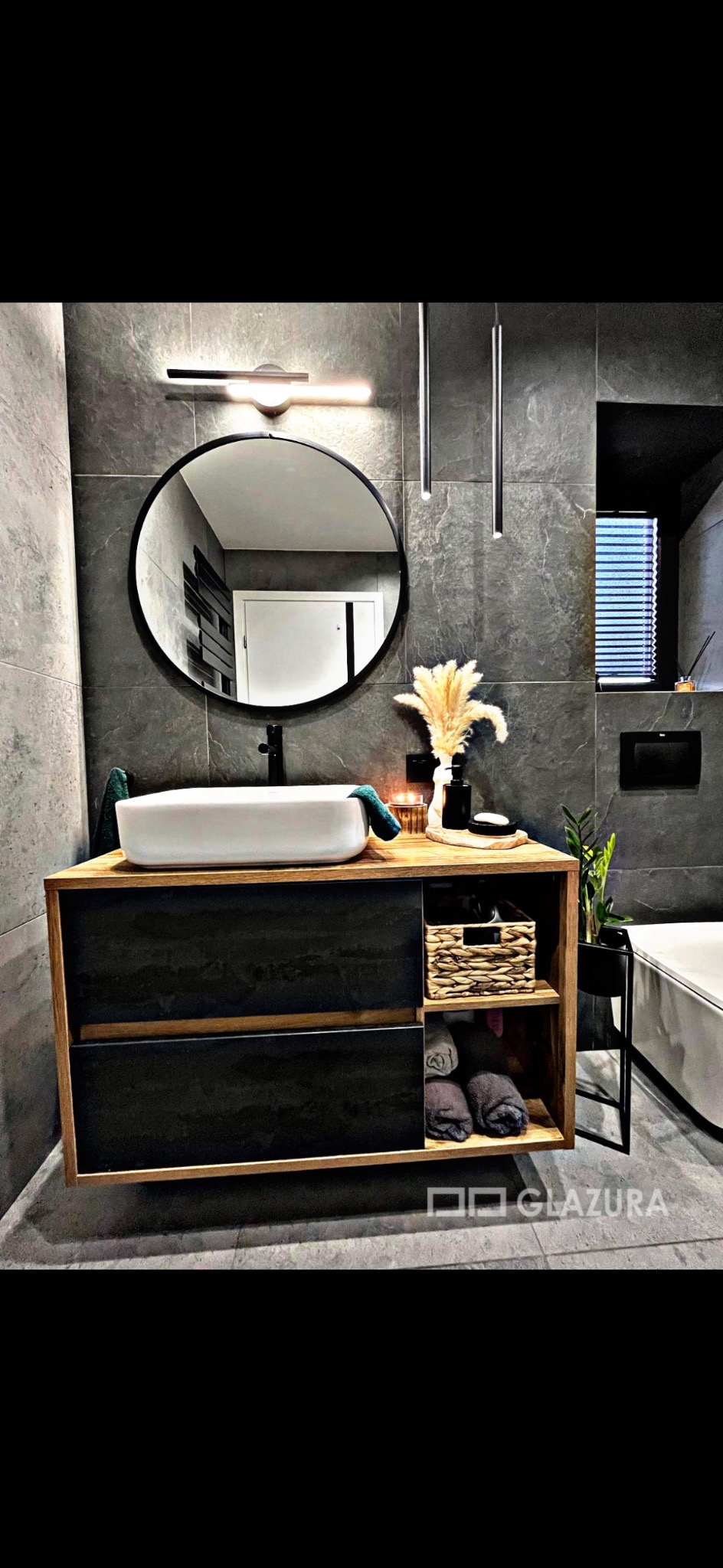 Stylowa łazienka z szarą glazurą, okrągłym lustrem i drewnianą szafką pod umywalką. Minimalistyczne oświetlenie i dekoracje dodają elegancji.