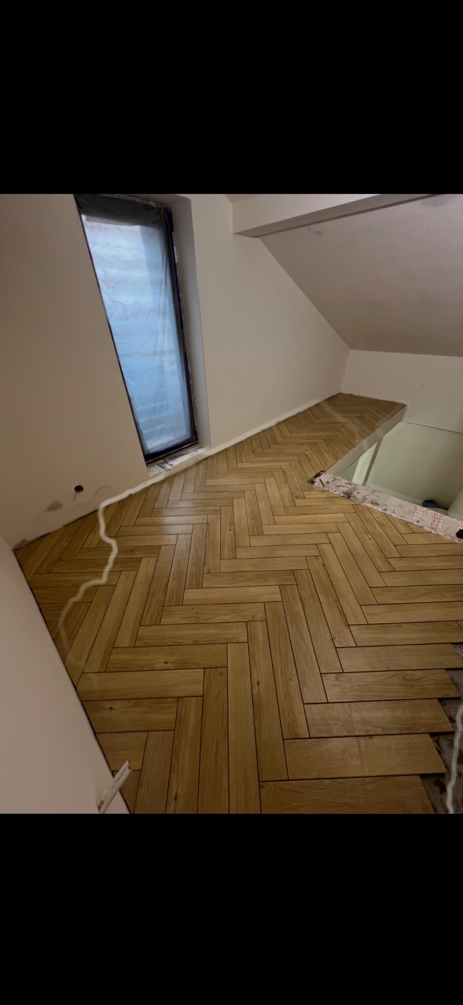 Ukończona podłoga z drewnopodobnych paneli ułożonych w jodełkę w pokoju na poddaszu z oknem dachowym. Częściowo widoczne niedokończone fragmenty podłogi.