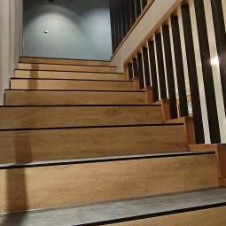 Schody wykończone panelami w odcieniach drewna, z czarnymi akcentami antypoślizgowymi. Widoczne drewniane barierki z czarnymi, pionowymi elementami. Ściana w tle w kolorze jasnego błękitu.