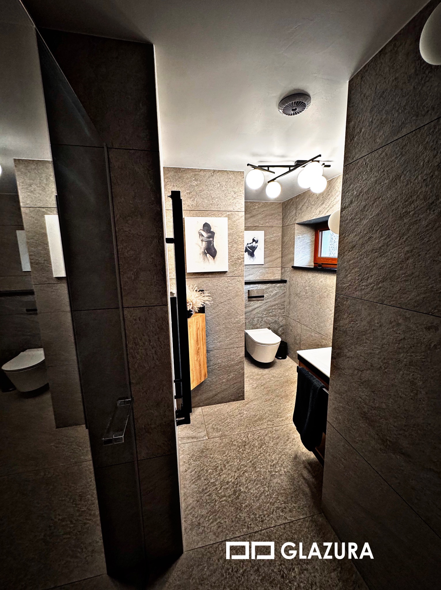 Stylowa łazienka z szarymi płytkami imitującymi kamień, oświetleniem sufitowym i dekoracyjnymi obrazami na ścianach. Widok zza szklanej kabiny prysznicowej.