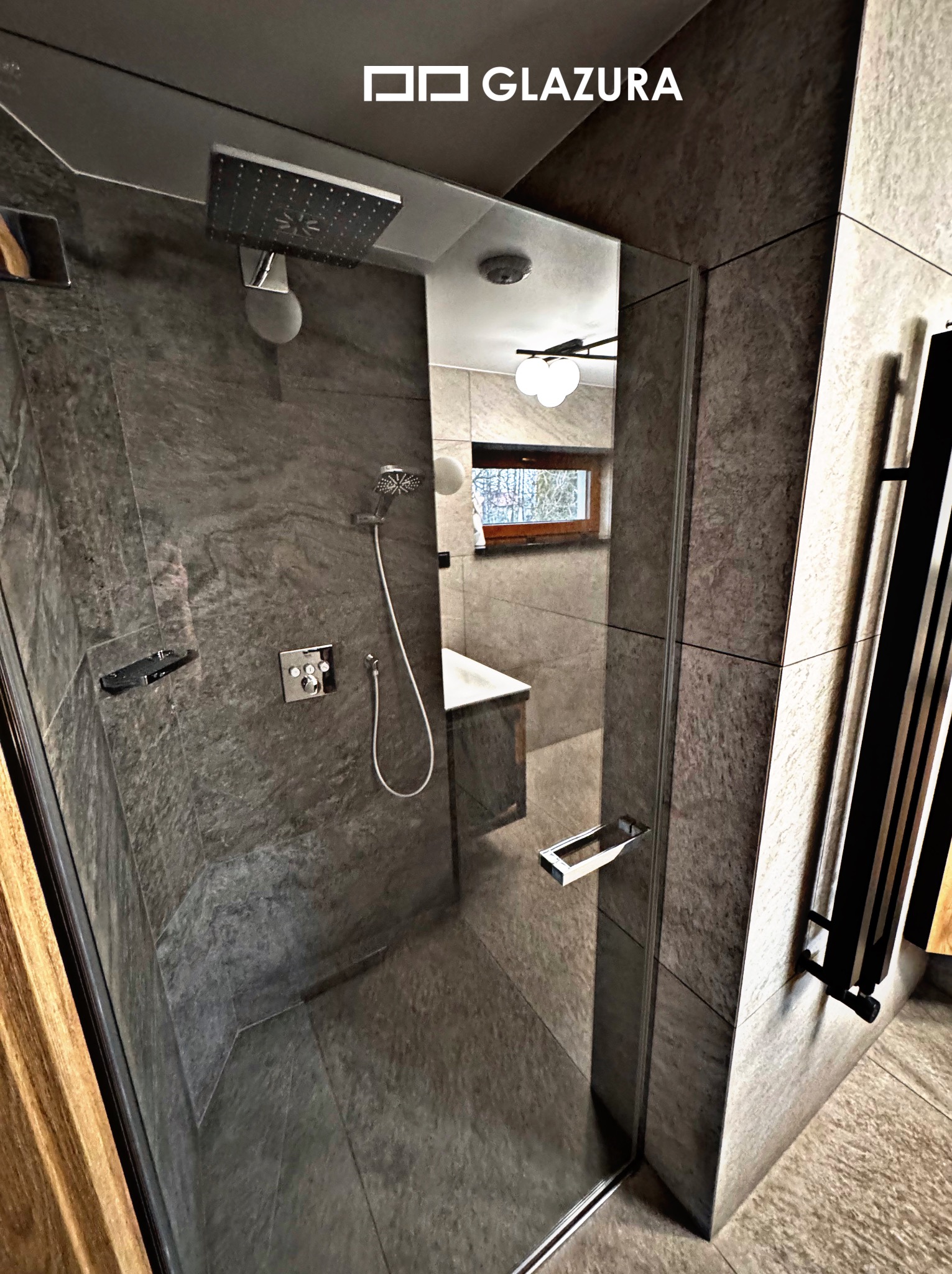 Nowoczesna kabina prysznicowa z szarymi płytkami imitującymi kamień, deszczownicą i szklaną ścianą. Widoczne detale wykończenia i grzejnik drabinkowy.