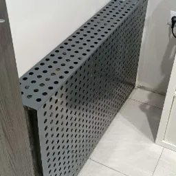 Szara, metalowa obudowa grzejnika z perforacją w postaci regularnych okręgów, stojąca przy białej ścianie i jasnej podłodze z płytek.