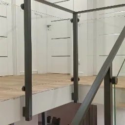 Szklana balustrada z czarnymi metalowymi słupkami i poręczą na antresoli, widok z dołu na stół i krzesła w jadalni, białe drzwi w tle.