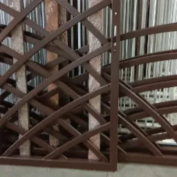 Brązowe metalowe panele z dekoracyjnym, krzyżowym wzorem, ustawione na szarej, betonowej podłodze. W tle widoczne pionowe, metalowe pręty.