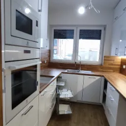 Nowoczesna kuchnia z białymi szafkami, drewnianym blatem i wysuwanymi koszami cargo, widok na okno z roletami.