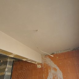 MARU - Świeżo położony, gładki, jasny tynk na suficie w surowym wnętrzu, widoczne cegły i wystający przewód elektryczny.