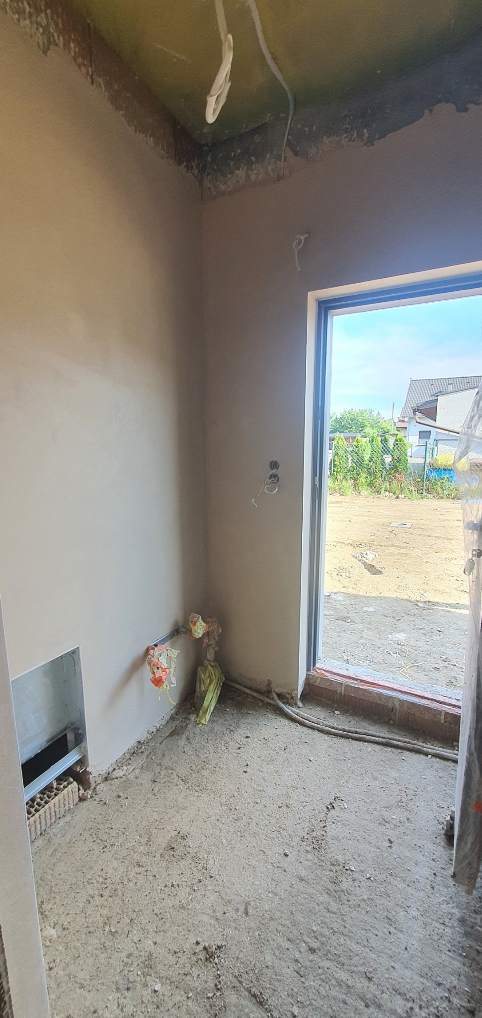 Wnętrze pomieszczenia po nałożeniu tynków maszynowych, widoczne wystające kable elektryczne i zabezpieczenia przy podłodze, przez otwarte drzwi widok na zewnątrz.