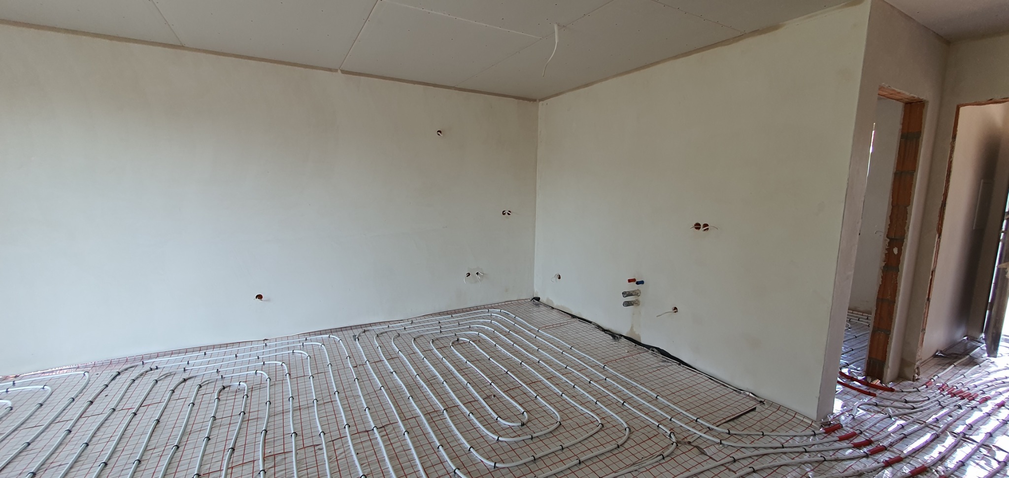 Surowe ściany po nałożeniu tynku maszynowego, widoczne otwory na instalacje elektryczne i hydrauliczne; na podłodze rozłożone rury ogrzewania podłogowego na macie grzewczej.