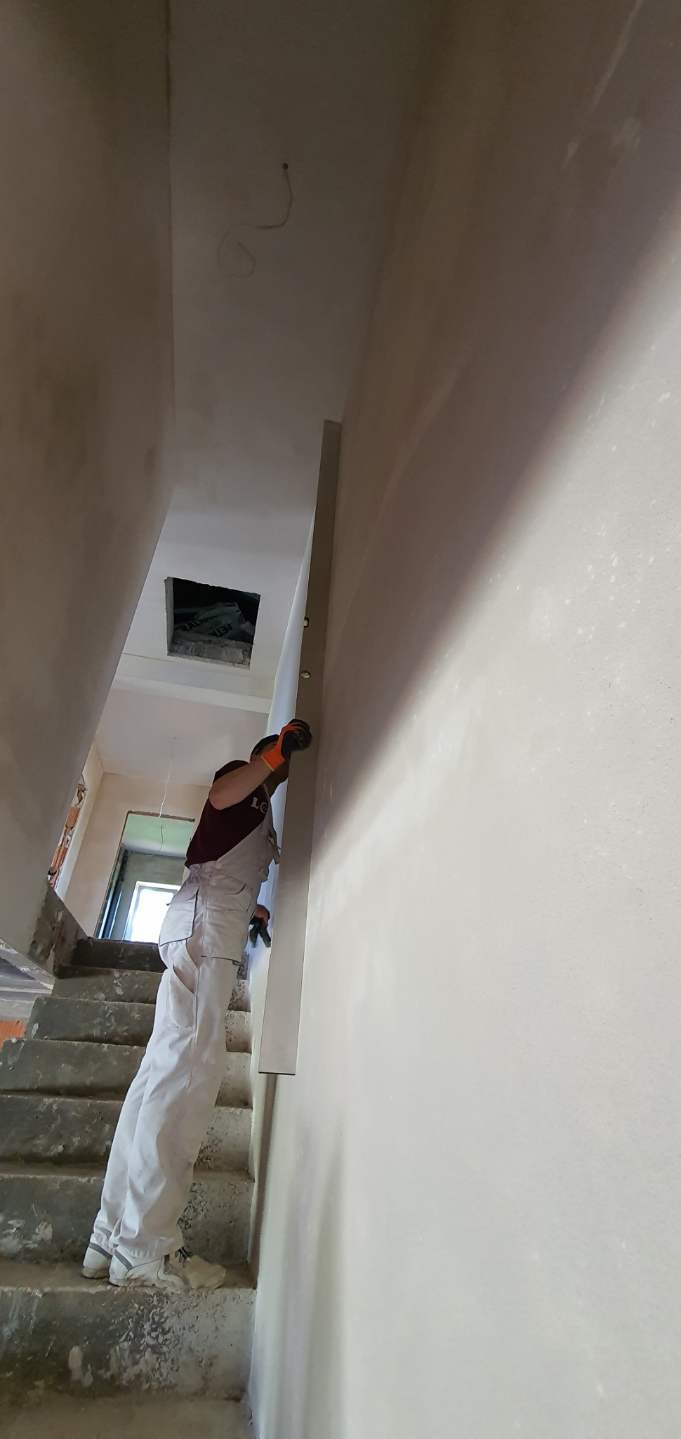 Pracownik w kombinezonie, na schodach, wyrównuje ścianę na klatce schodowej za pomocą szpachli i poziomicy.