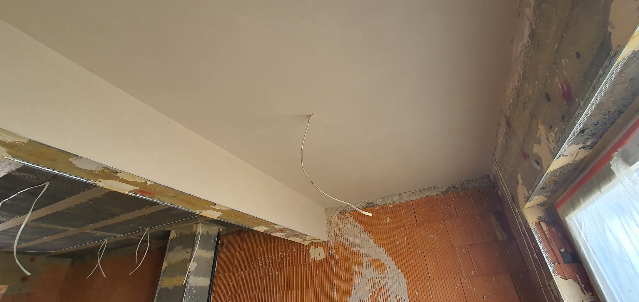 Świeżo położony, gładki, jasny tynk na suficie w surowym wnętrzu, widoczne cegły i wystający przewód elektryczny.