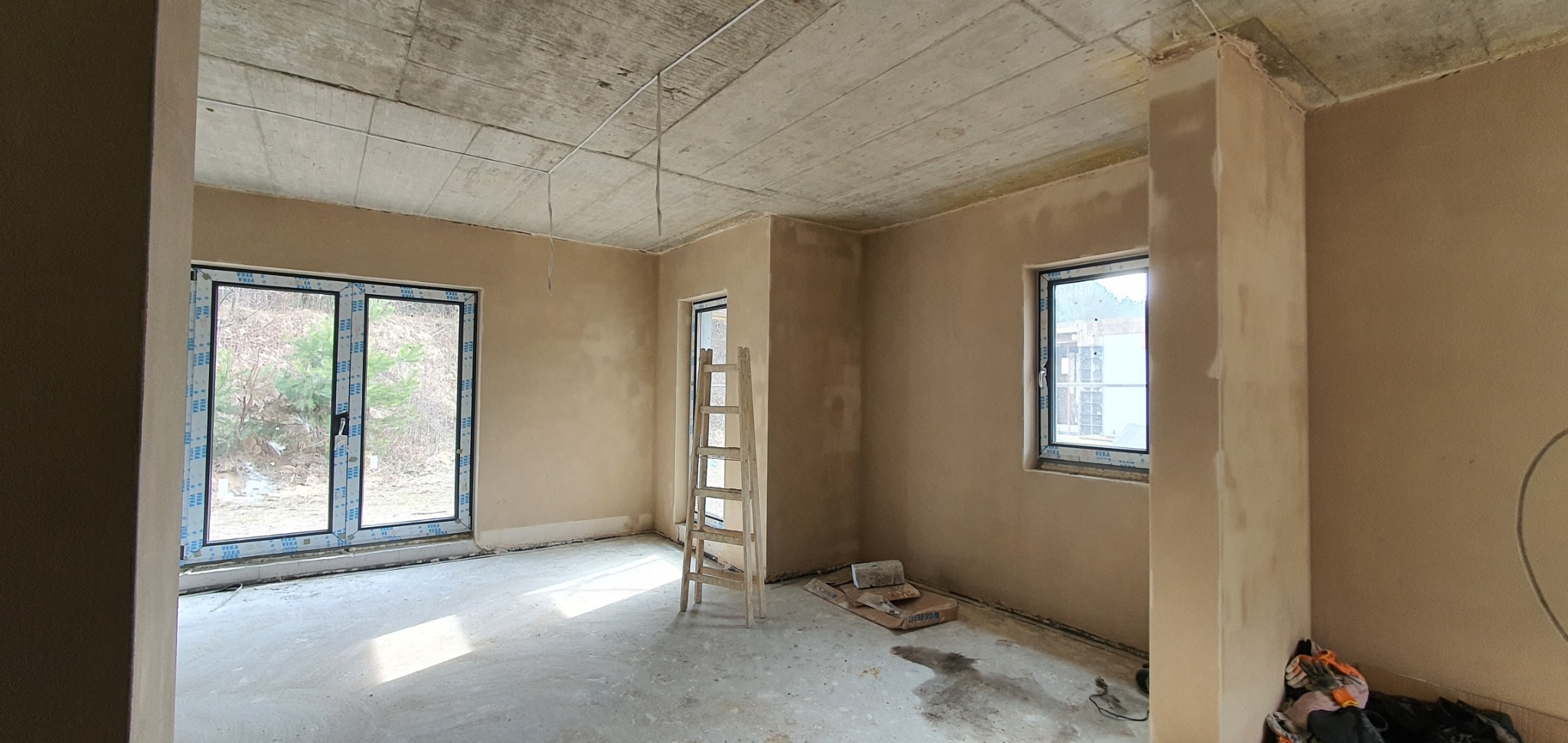 Wnętrze po nałożeniu tynków: surowy betonowy strop, ściany pokryte świeżym tynkiem, drewniana drabina oparta o ścianę, okna z widokiem na zieleń.