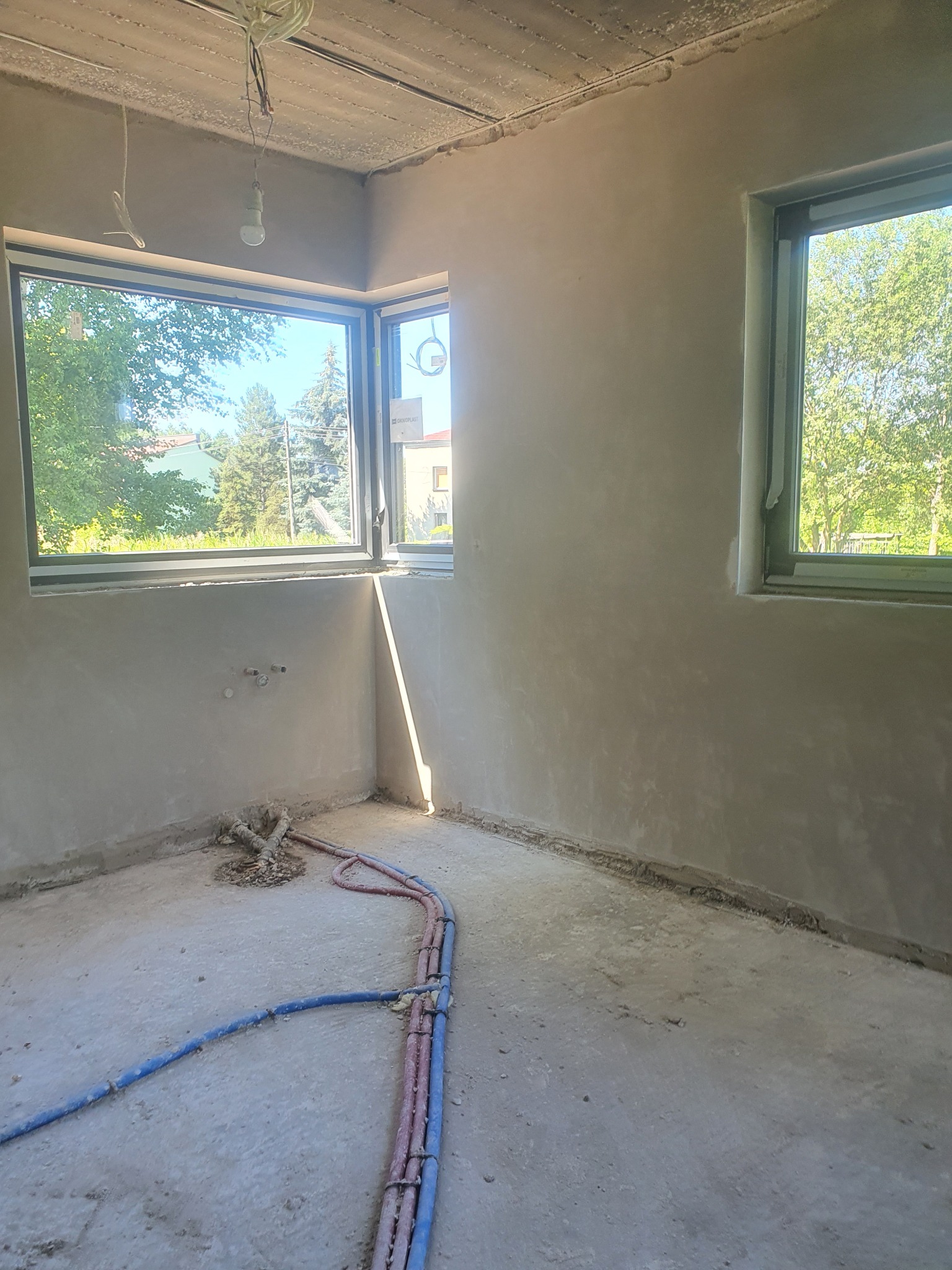 Wnętrze pokoju po tynkowaniu maszynowym ścian i sufitu, widoczne rury instalacji wodnej na podłodze, dwa okna z widokiem na zieleń na zewnątrz.