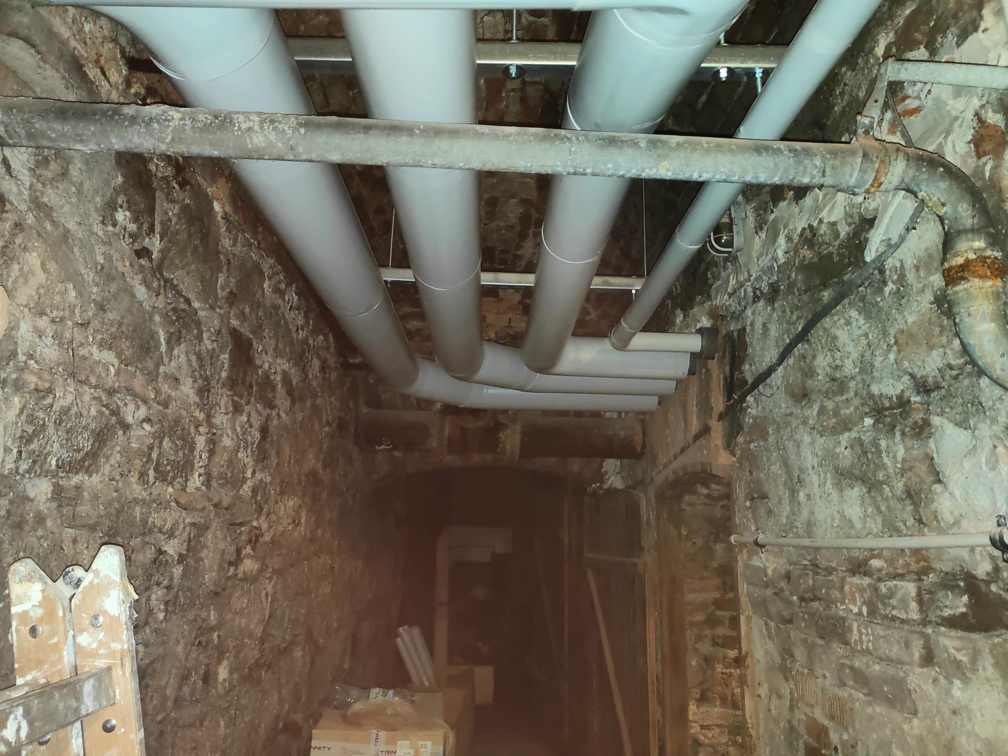 Piwnica z odsłoniętą cegłą, w której zamontowano nowe rury CO/CWU, wsparte na metalowych profilach, obok starej, zardzewiałej instalacji.