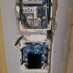 Volt Customs -Maksym Ruban - Dwa otwarte panele elektryczne w ścianie w trakcie montażu, z widocznymi przewodami, bezpiecznikami i urządzeniami, ściana surowa, widoczne ślady po kuciu.