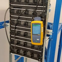 Volt Customs -Maksym Ruban - Panel krosowniczy w szafie rack, testowany żółtym miernikiem kabli, z widocznymi niebieskimi kablami sieciowymi.