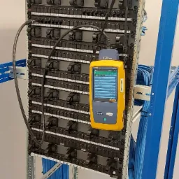 Panel krosowniczy w szafie rack, testowany żółtym miernikiem kabli, z widocznymi niebieskimi kablami sieciowymi.