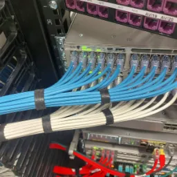 Szafa rack z uporządkowanymi kablami sieciowymi: niebieskie i białe przewody spięte rzepami, podłączone do przełączników Cisco z diodami LED.