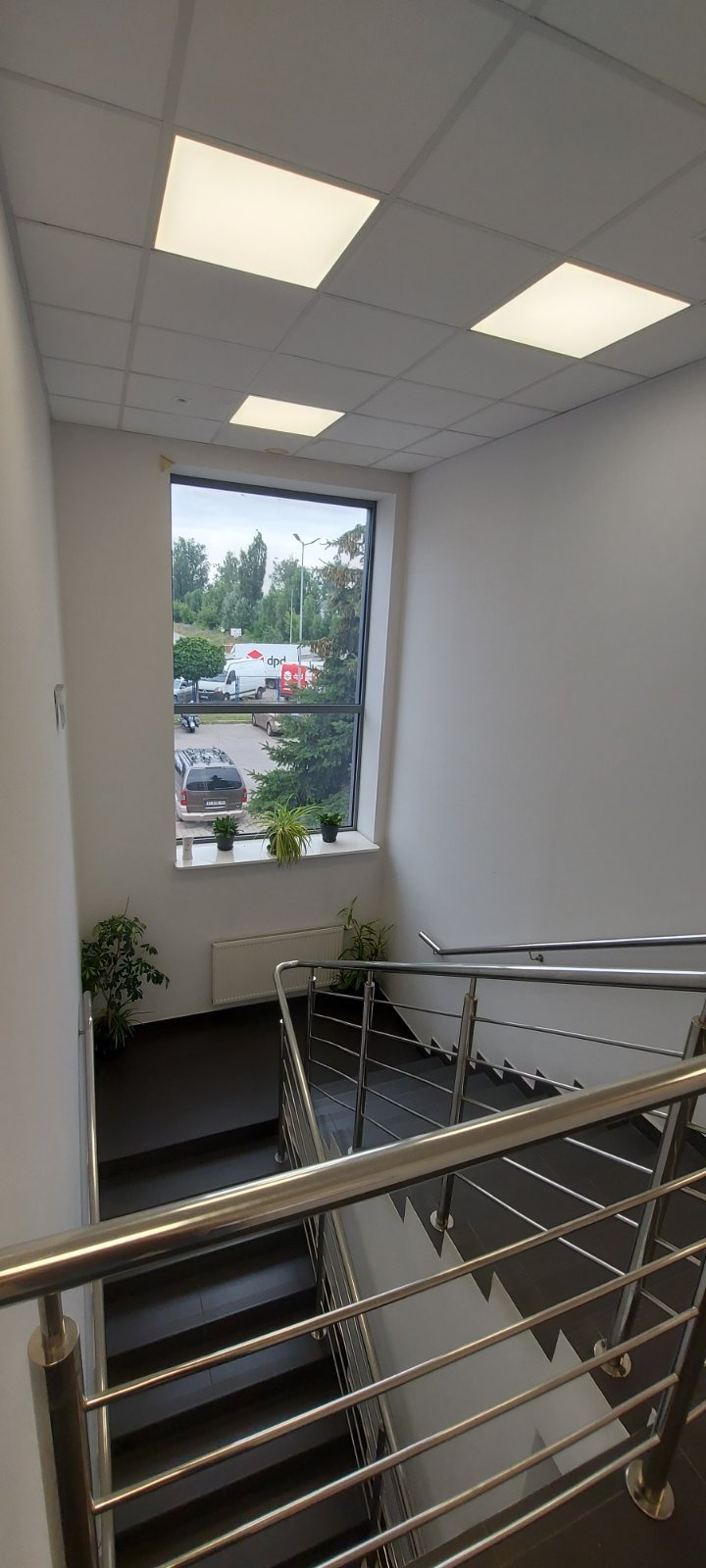 Wnętrze klatki schodowej z metalową balustradą, widok na okno z roślinami doniczkowymi i zaparkowane samochody, oświetlenie sufitowe rastrowe.
