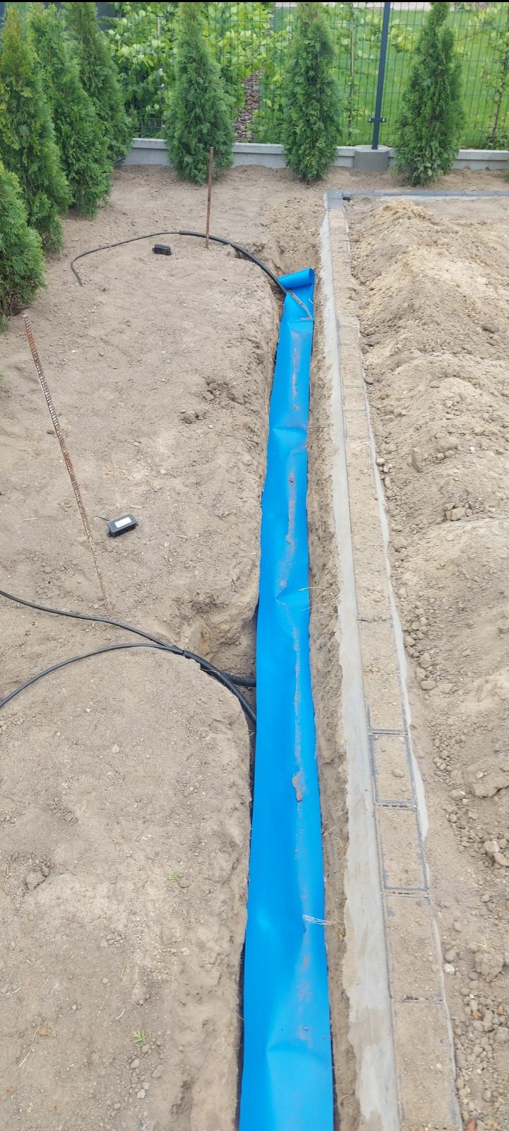 Wykop w ziemi wyłożony niebieską folią ochronną, obok ułożony chodnik z kostki, widoczne kable elektryczne w czarnej rurze karbowanej.