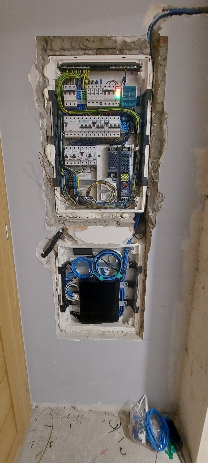 Dwa otwarte panele elektryczne w ścianie w trakcie montażu, z widocznymi przewodami, bezpiecznikami i urządzeniami, ściana surowa, widoczne ślady po kuciu.