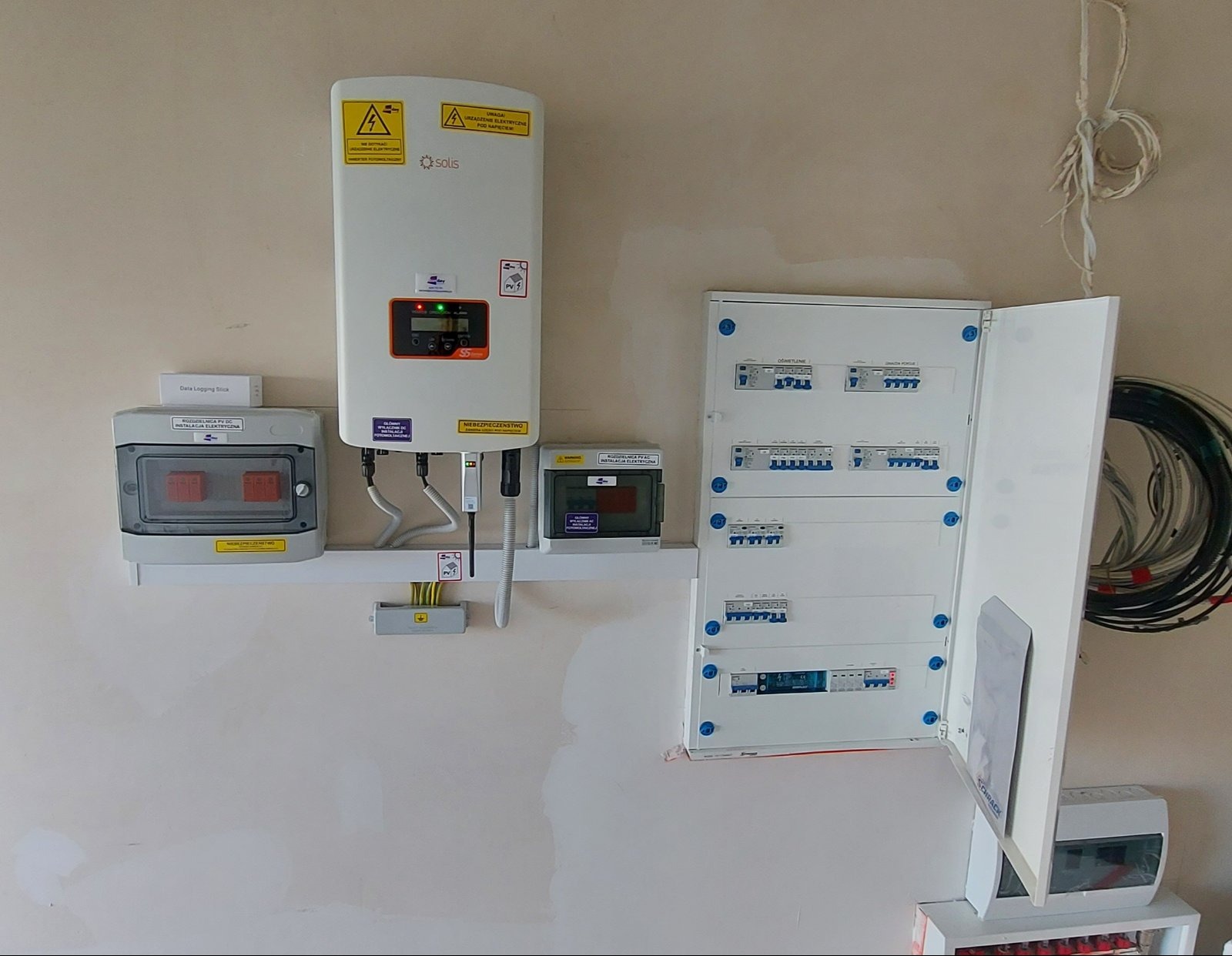 Rozdzielnia elektryczna z falownikiem solis, zabezpieczenia i przewody na ścianie w pomieszczeniu o jasnych ścianach.