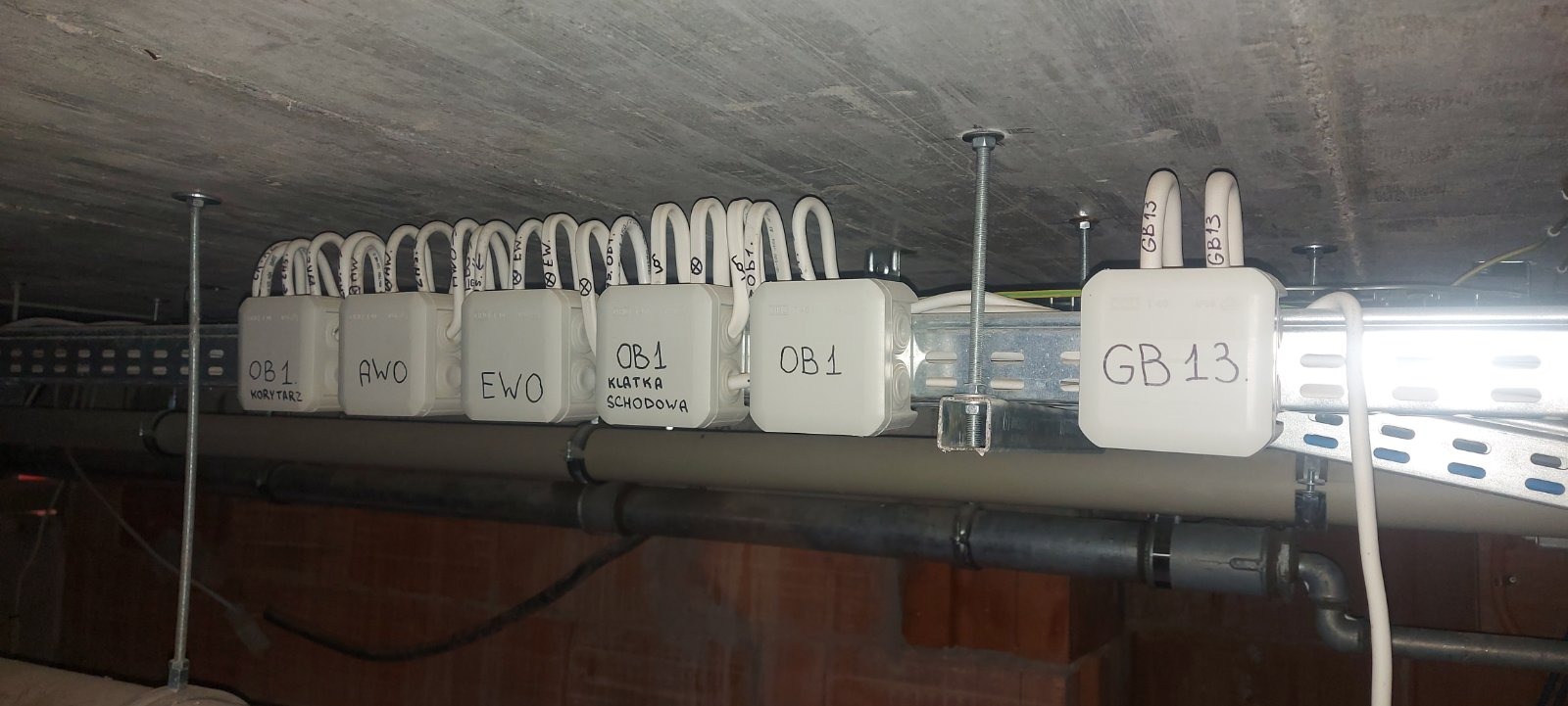 Rząd białych puszek elektrycznych z opisami (OB1, AWO, EWO, KLATKA SCHODOWA, GB13) przymocowanych do metalowej szyny pod sufitem, widoczne przewody i rury instalacyjne.